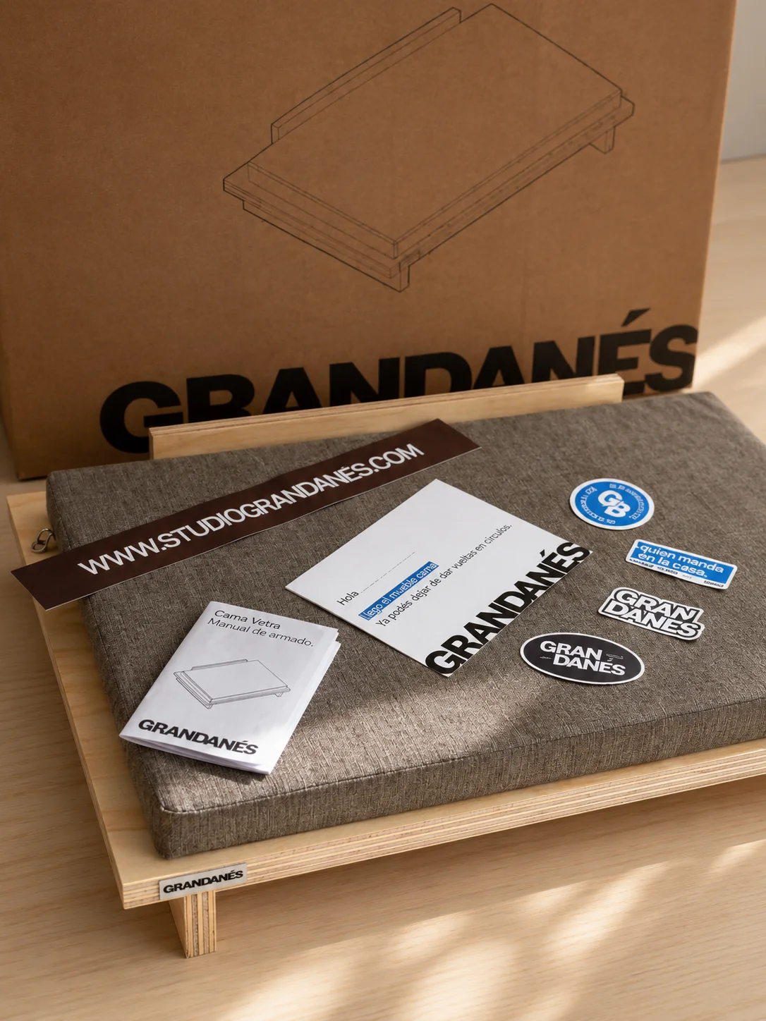 The Vetra bed with its cushion, a sticker sheet and cards, in front of its cardboard box with the GRANDANÉS logo.